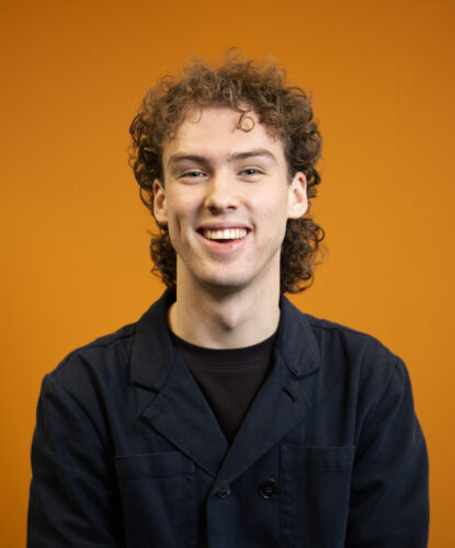 Portretfoto van een jonge persoon met krullend haar en een brede glimlach, gefotografeerd tegen een feloranje achtergrond. De persoon draagt een donkerblauw jasje over een zwart shirt en kijkt recht in de camera.