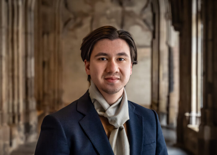 Portretfoto van een man met donker haar in een donkerblauwe blazer en okergele trui met een lichtgrijze sjaal, poserend voor de onscherpe achtergrond van een historische zuilengang.